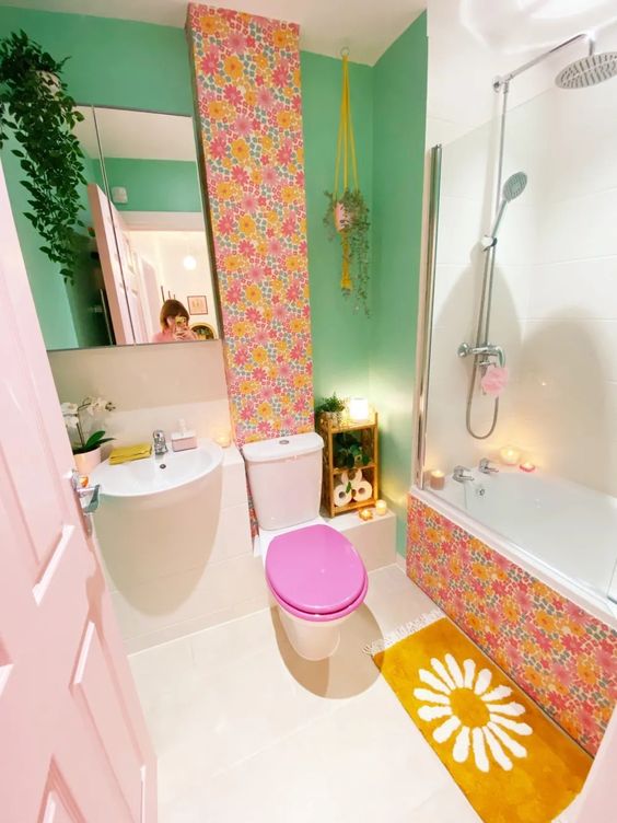 A bathroom with dopamine decor, a green wall, wallpaper accents, a mustard rug, green plants and a blush door