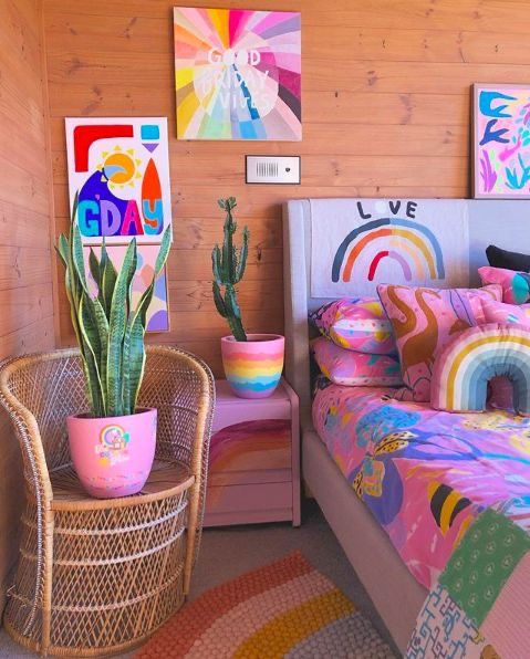 A bedroom with dopamine decor and a bed with extra statement linens, pink nightstands, potted plants and colorful artwork