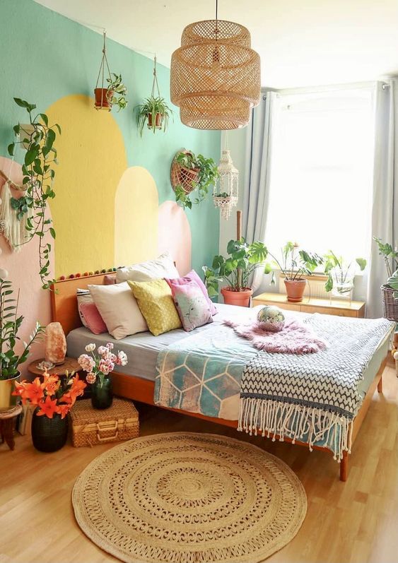 A bedroom in dopamine decor with a cool and colorful accent wall, a bed with pastel linens, flowers, a pendant lamp and lots of greenery