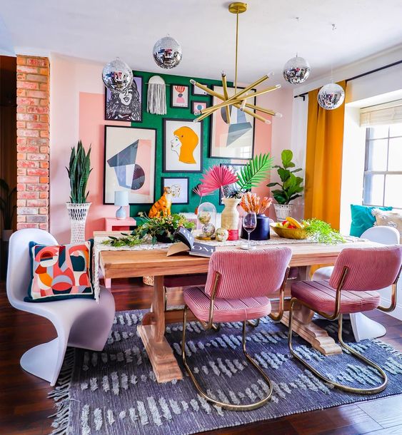 A dopamine decor dining room with a green accent wall and a statement gallery wall, table, pink chairs, colorful pillows and lots of greenery