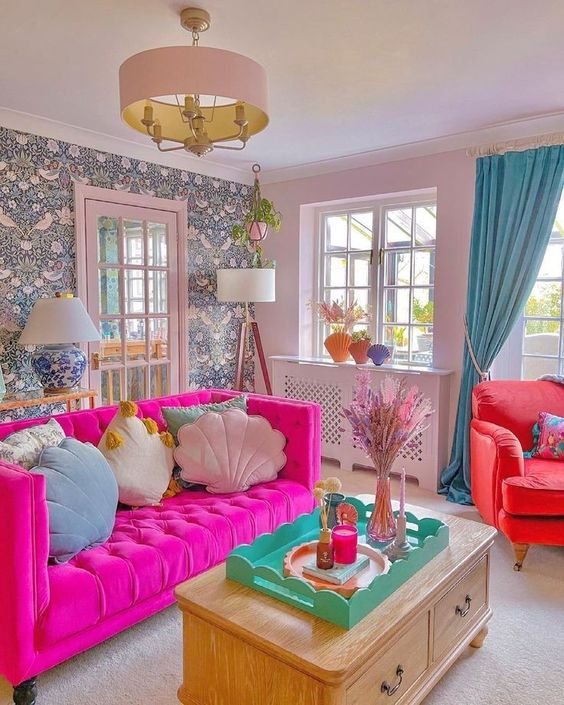 A living room in dopamine decor with a wallpaper wall, a pink sofa with pastel pillows, a coral armchair and blue curtains