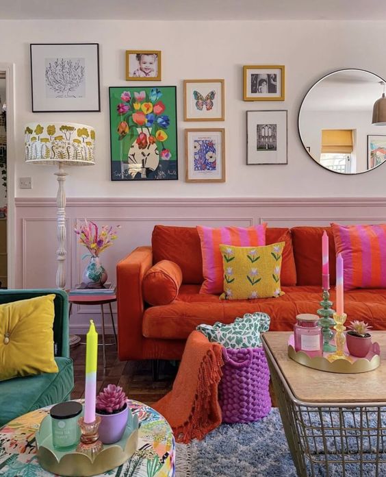 A living room in dopamine decor with blush paneling, an orange sofa and colorful pillows, a hot pink basket and a pretty gallery wall