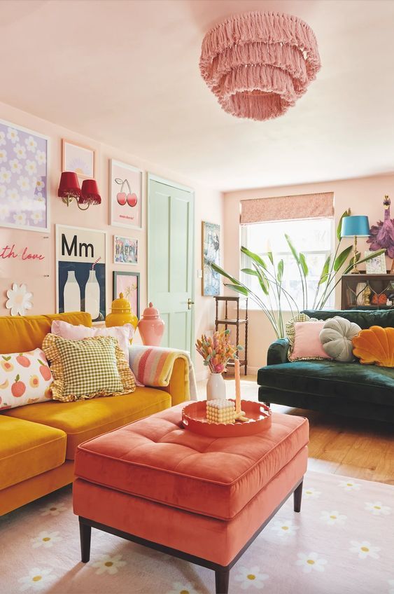 A living room in dopamine decor with blush walls, a marigold sofa, a coral stool, a dark green chair, a gallery wall and a pink chandelier