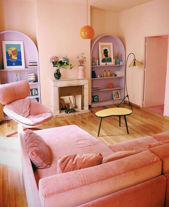 A living room in dopamine decor with blush walls and ceiling, a pink sofa and chair, purple shelves and a hanging lamp