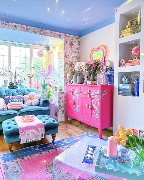 A dopamine decor living room with floral wallpaper, a navy sofa and ottoman, pastel pillows, a hot pink credenza, statement decor, and blooms