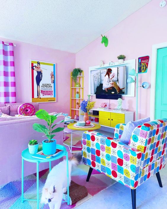 A living room in dopamine decor with light pink walls, a purple sofa with pillows, a colorful printed chair, a turquoise side table and eye-catching decor