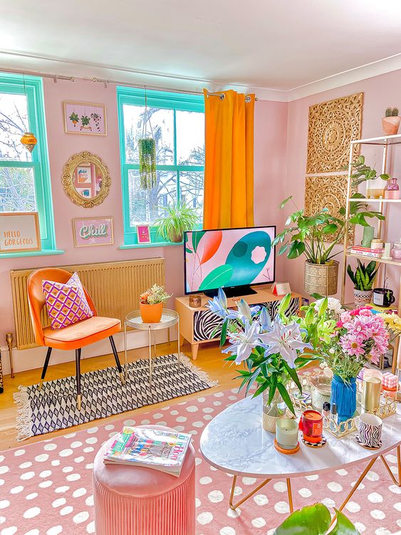 A dopamine decor living room with pink walls, turquoise windows, an orange chair, a pink printed rug, a pink stool and some flowers and greenery