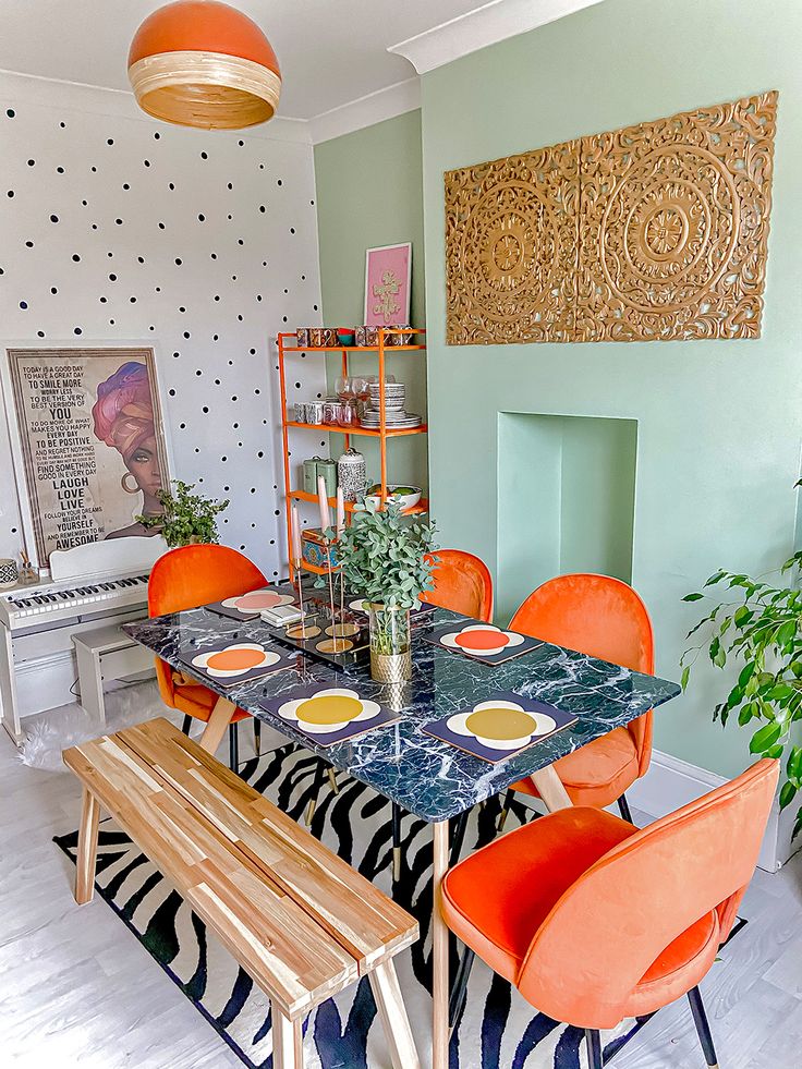 a dopamine dining area with a mint and polka dot accent wall, marble table, orange chairs and shelf, lots of greenery