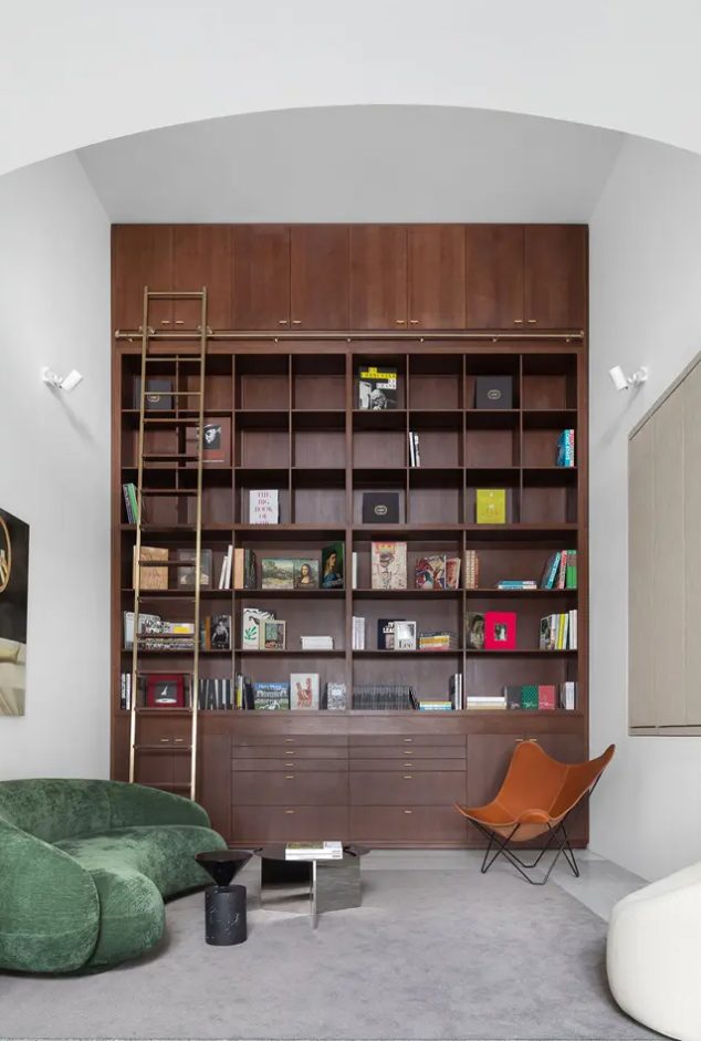 a home library with a large bookcase and ladder, a green curved low sofa, a cream chair and a leather chair, and side tables