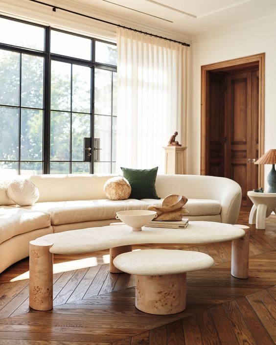 a light-filled living room with parquet floors, a curved neutral sofa, a coffee table, a stool and a few lamps