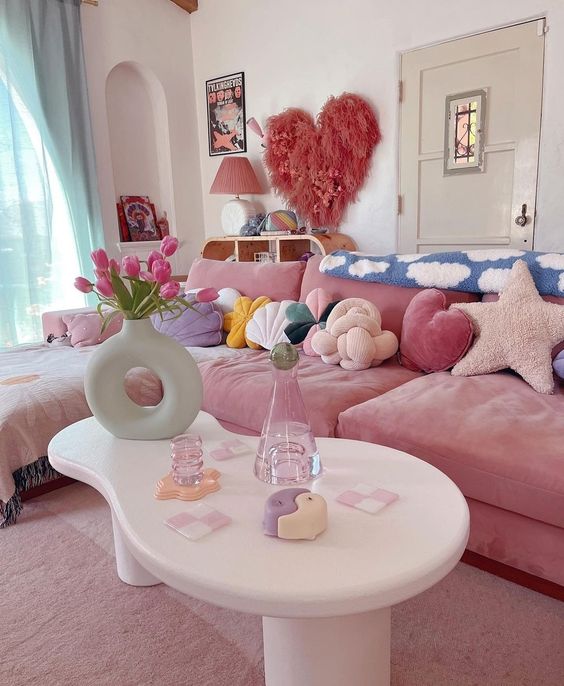 a living room with a pink sofa, colorful pillows, turquoise curtains, a heart and some art and a table with pretty decorations
