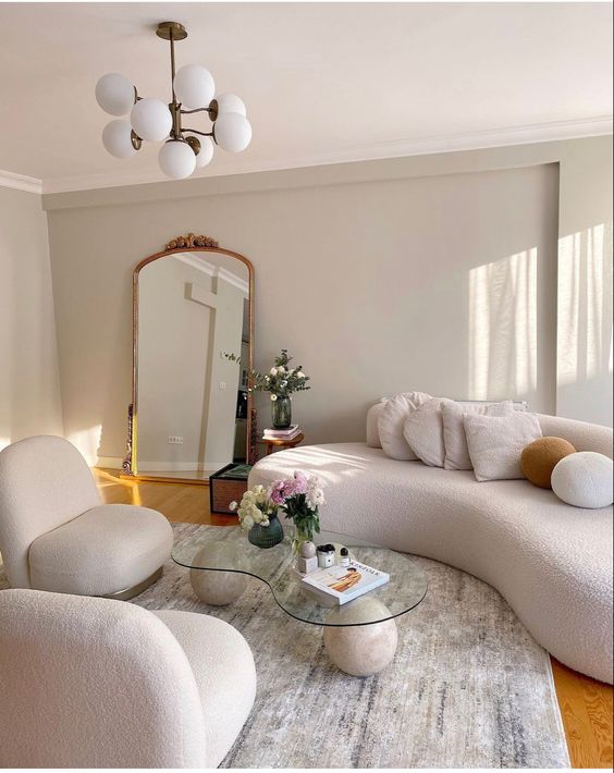 a pretty, neutral living room with cream curved bouclé furniture, a matching glass coffee table and a gold-framed mirror
