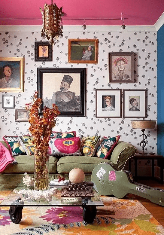 a whimsical living room with a pink blanket, printed wallpaper, a green sofa, a gallery wall, a glass coffee table and a statement rug