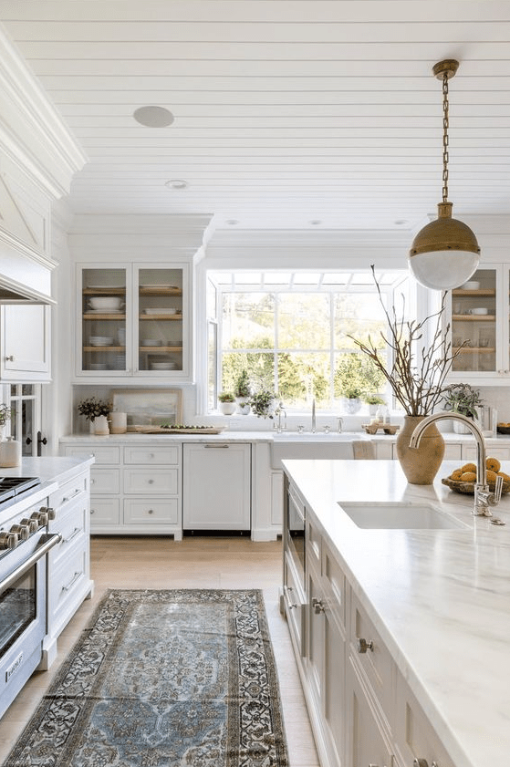 a serene white kitchen with marquetry and glass cabinets, a white stone backsplash and countertops, and brass pendant lamps
