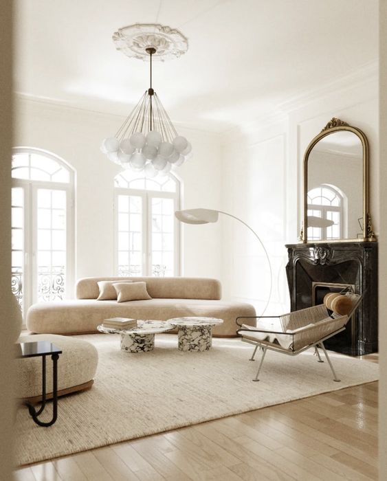 an elegant neutral living room with a black marble fireplace, neutral curved sofa, chair, marble coffee tables and a bubble chandelier