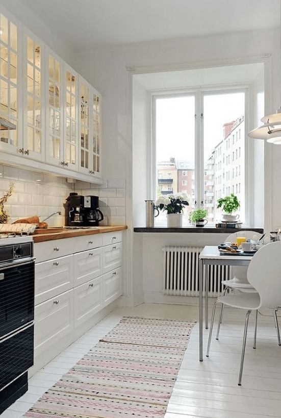 an inviting white Scandinavian kitchen with glass cabinets, white tiles, butcher block countertops and a small dining area