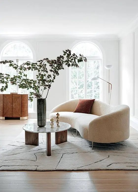 an airy Scandinavian living room with arched windows, a cream curved sofa, a chic sideboard and a three-legged round table and a floor lamp