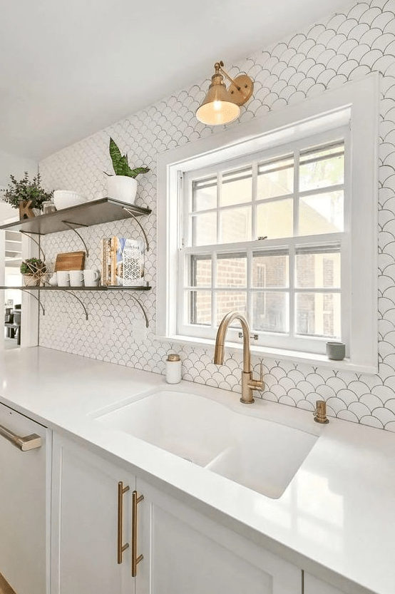 an airy white kitchen with shaker cabinets, a white fish scale tile backsplash, open shelving, and brass fixtures and lamps
