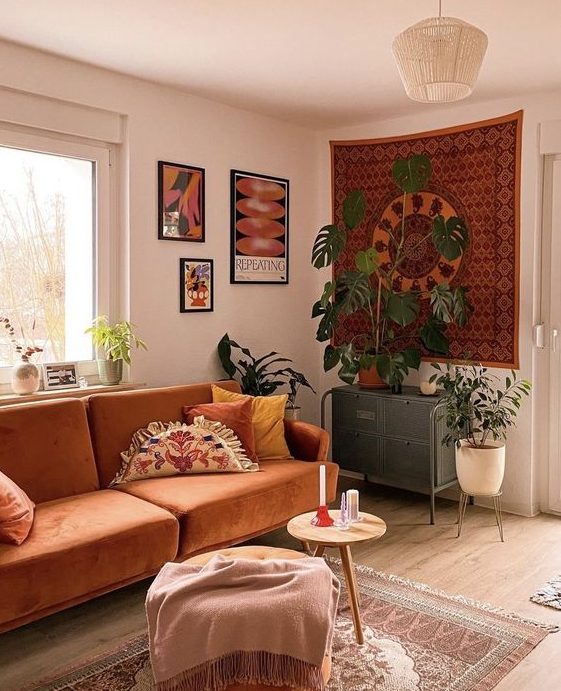 A boho living room in earth tones with some artwork, a rust-colored sofa, stools and ottomans, and potted plants