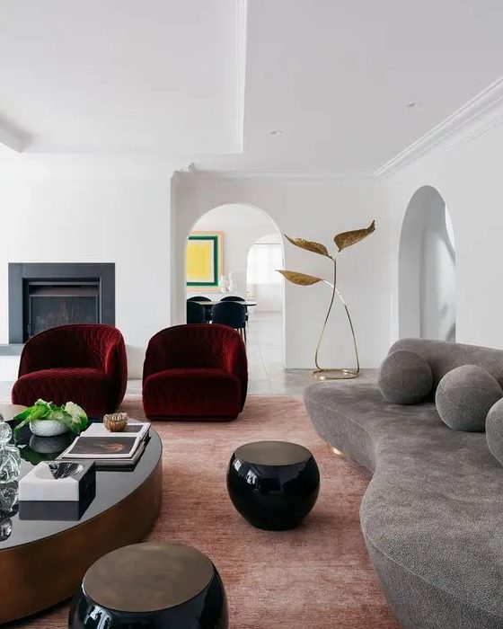 an exquisite living room with a fireplace, a gray curved sofa with cushions, burgundy curved chairs and rounded tables