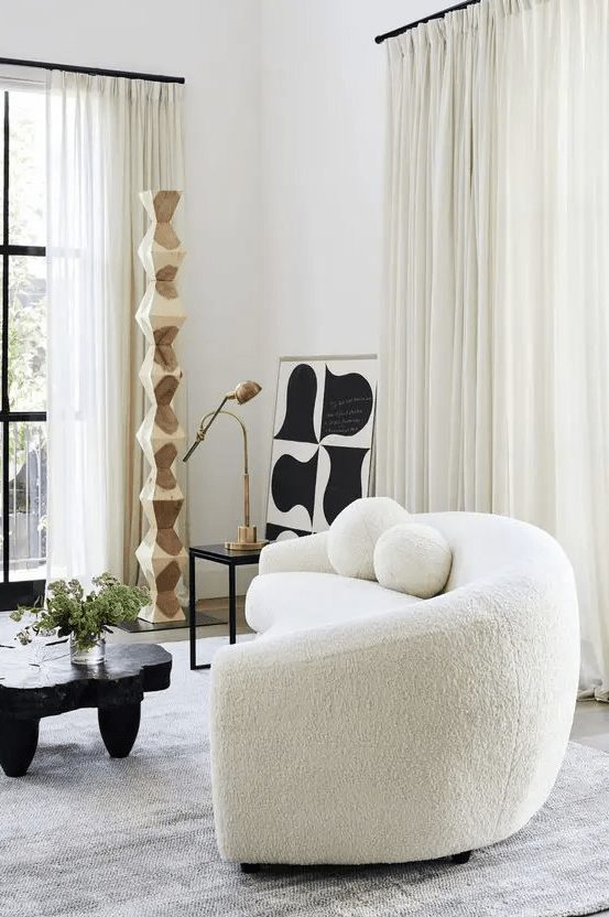 A striking room with a white curved bouclé sofa, a black tree slice table, a side table with a lamp and some abstract art