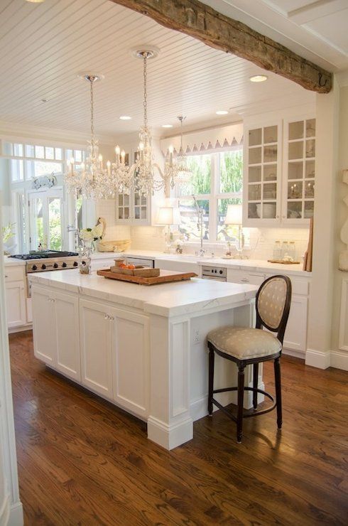 a white farmhouse kitchen with shaker and glass cabinets, a wood beam, crystal chandeliers and a vintage chair