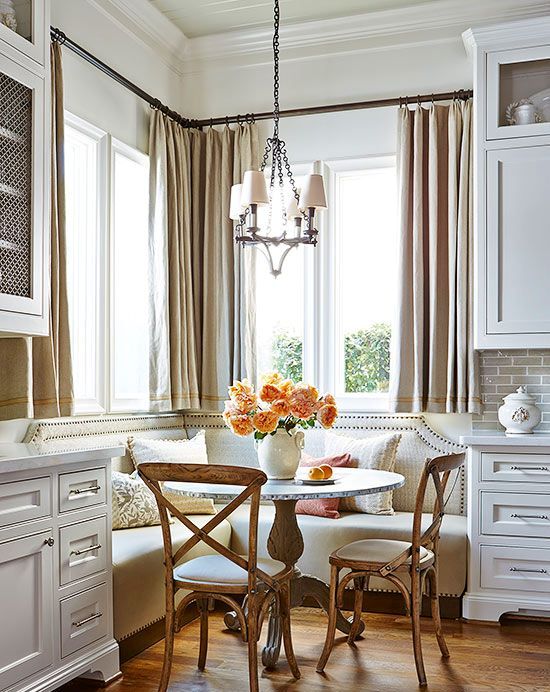 a cozy and elegant country-style breakfast nook with a built-in upholstered bench, a table and stained chairs, a hanging lamp and some flowers