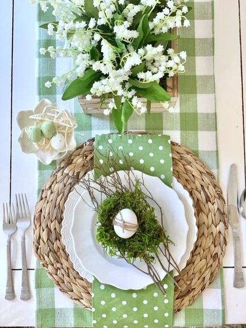 a green rustic Easter tablescape with a plaid runner, polka dot napkin, woven placemat, lily of the valley centerpiece and a moss nest with an egg
