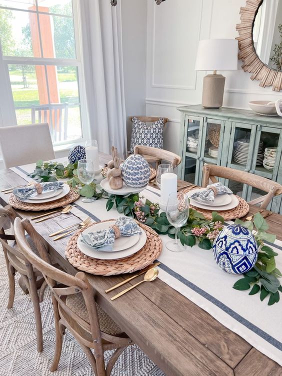 A pretty farmhouse Easter tablescape with a neutral runner, greenery, blue printed eggs and napkins, woven placemats and gold cutlery