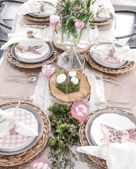 a beautiful rustic Easter tablescape with woven placemats, greenery, moss, checkered plates, terrariums with moss and eggs and pink tulips