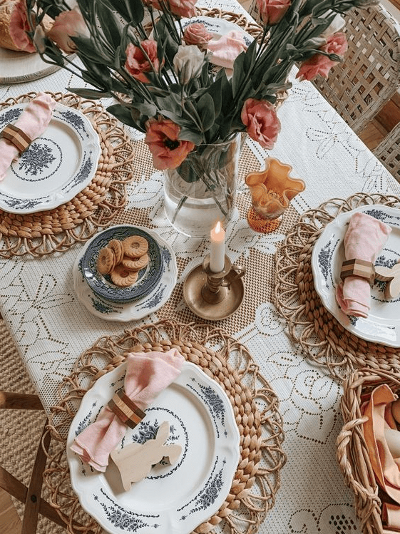 A modern farmhouse style tablescape featuring a doily tablecloth, woven tablecloths, printed plates, pink napkins, fresh flowers and candles