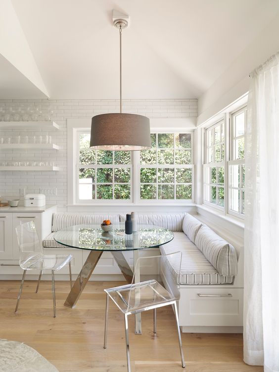 a neutral, country-style breakfast nook with large corner banquet seating, a glass table and acrylic chairs, and a hanging lamp