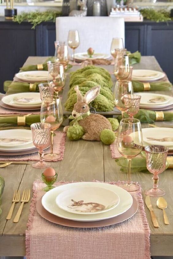 A pastel Easter tablescape with pink runners, green napkins, moss, a bunny and pink glasses is beautiful