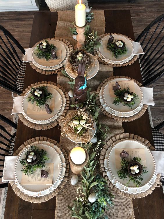 A rustic Easter table setting with a burlap runner and placemats, white china, nests with eggs, greenery and candles is a cool idea for a party