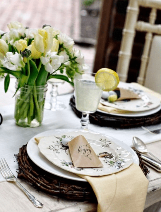 a rustic Easter table with a white and yellow tulip centerpiece, vine-patterned printed porcelain placemats, and chic cutlery and elegant glassware