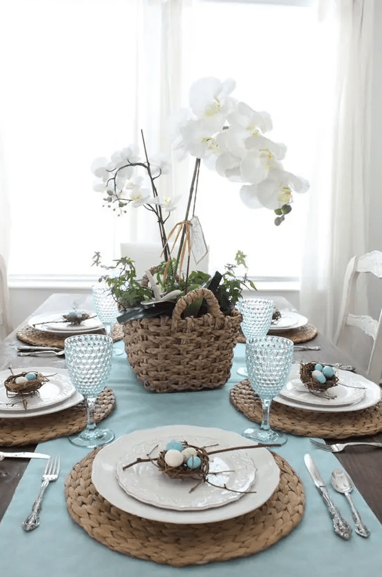 a rustic Easter tablescape with a blue runner, woven placemats, a basket of greenery and white orchids, and mini nests of eggs