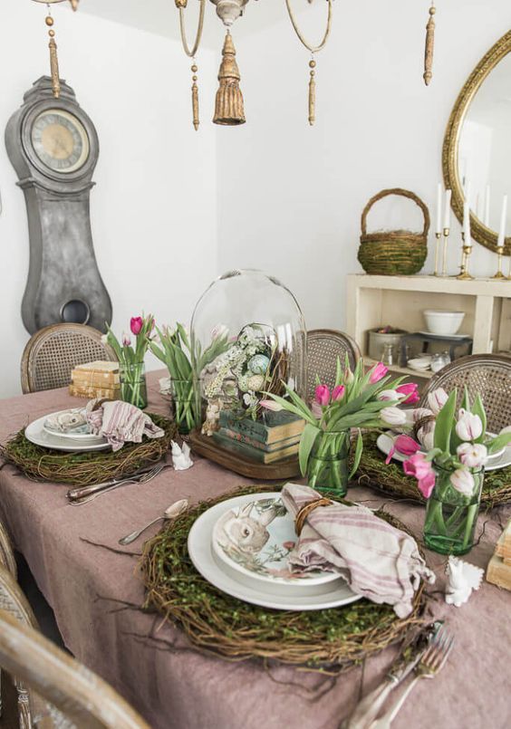 A rustic Easter tablescape with an egg cloche, vine and moss placemats, pink and white tulips and greenery, and stacked books is wow